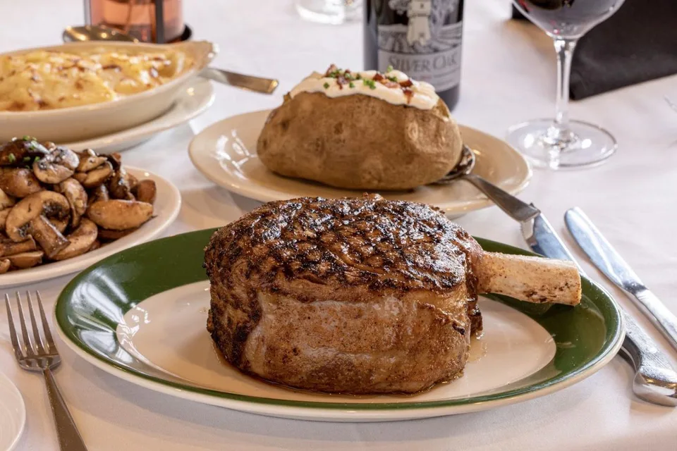 A bone-in steak sits on a green-rimmed plate with a baked potato and sautéed mushrooms.