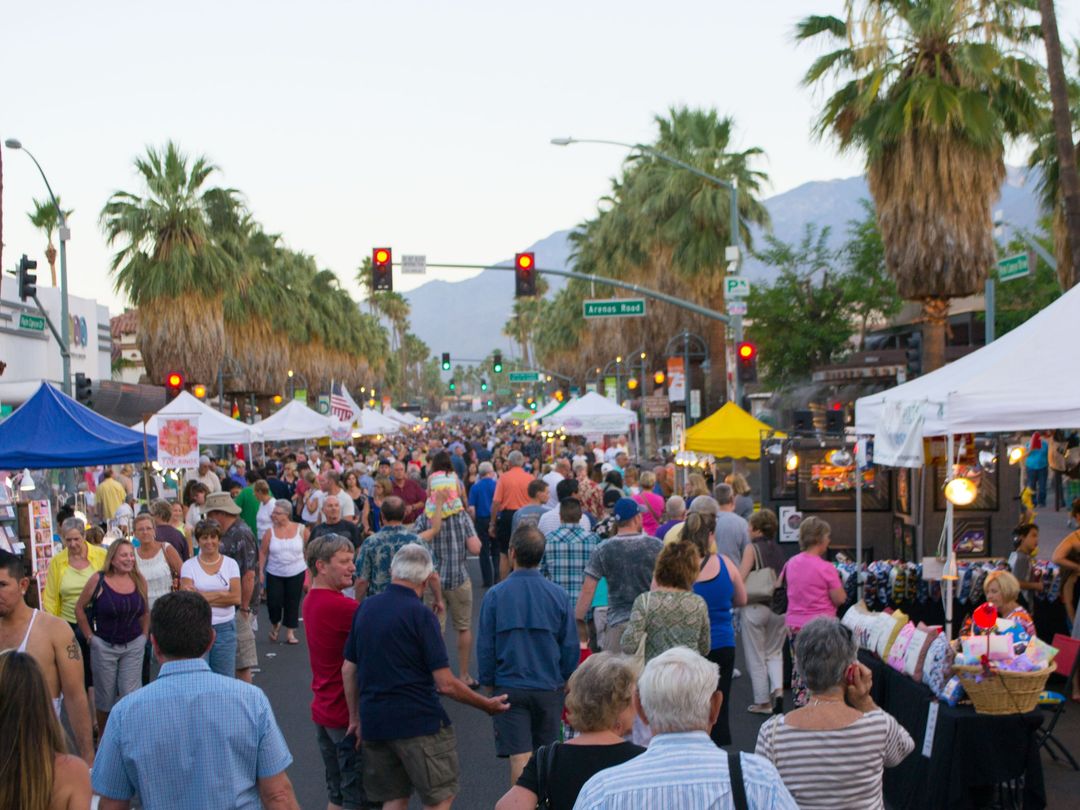 Palm Springs VillageFest fills Palm Canyon Drive with vendor tents and an evening crowd beneath palms and the San Jacinto Mountains.