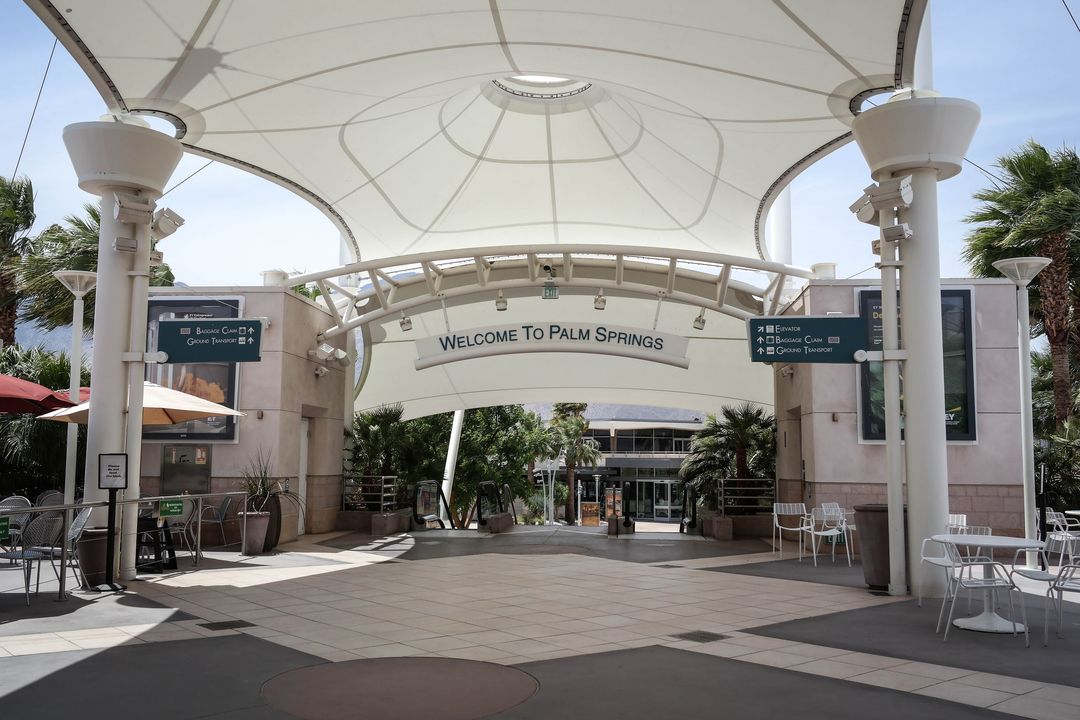 Open-air concourse at Palm Springs International Airport beneath a white tensile canopy and a Welcome to Palm Springs arch.