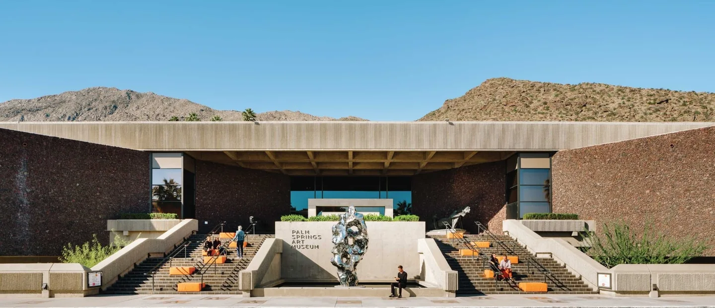 The Palm Springs Art Museum entrance, broad steps, and a reflective metal sculpture sit beneath the San Jacinto Mountains.
