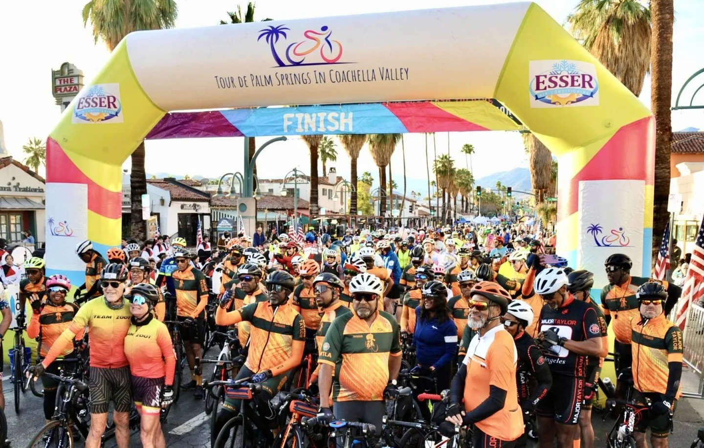 A large group of cyclists gathers beneath the Tour de Palm Springs finish arch on a palm-lined downtown street.