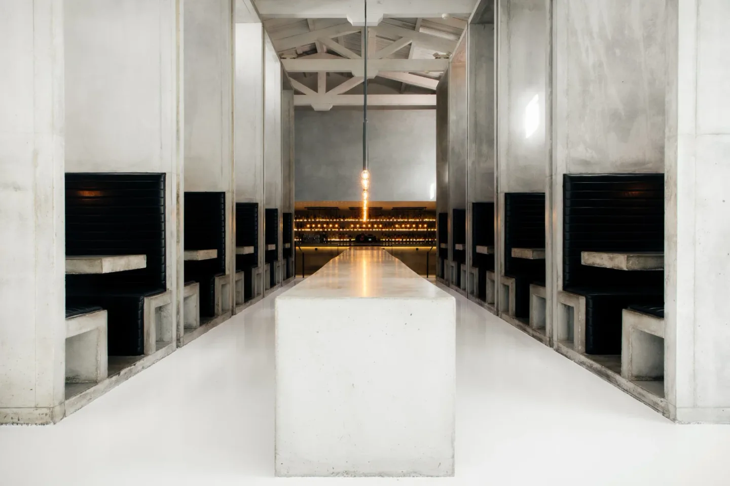 A long white communal table leads between concrete booths toward the illuminated bar at Workshop.
